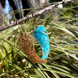 May include: A pair of handmade earrings with a wing design. One earring is gold-colored with a hammered texture, and the other is turquoise and blue. The earrings are hanging from a branch against a background of green and white foliage.