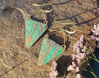 May include: A pair of handmade earrings with a triangular design. The earrings feature a green and copper-colored pattern and are suspended from gold-colored hooks. The image also includes the text "@anaisisleshandmadejewelry".