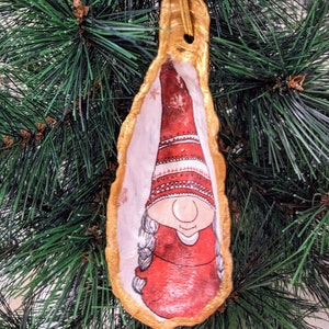 May include: A handmade Christmas ornament featuring a gnome with a red hat and white trim, painted on a gold-painted oyster shell. The ornament is hanging from a gold string.