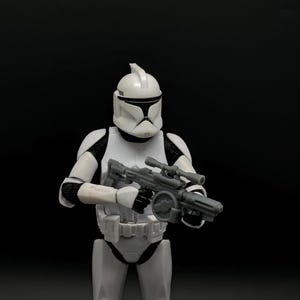 May include: A white and black action figure of a clone trooper holding a gray blaster rifle. The figure is wearing a white helmet and armor with black accents. The background is solid black.