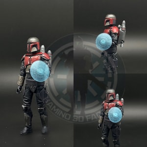 May include: A red and black action figure of a Mandalorian warrior holding a blue energy shield. The figure has a helmet with a visor and a jetpack. The figure is posed in a fighting stance.
