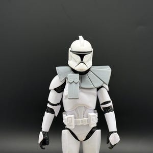 May include: A white and gray action figure of a clone trooper from Star Wars. The figure has a white helmet with black accents, gray shoulder armor, and a white and gray uniform. The figure is standing against a dark gray background.