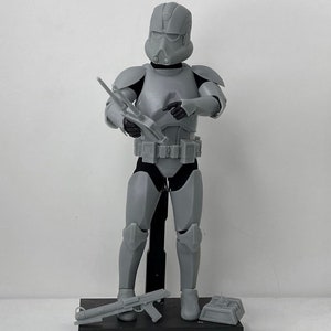 Può includere: Una action figure di plastica grigia di un clone trooper di Star Wars che tiene un fucile blaster. La figura è in piedi su una base nera con un supporto in plastica grigia.