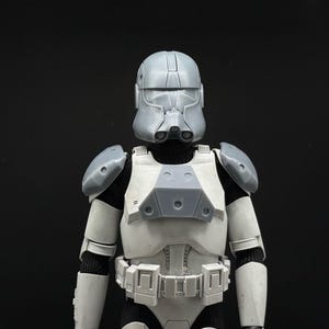 May include: A gray and white action figure of a futuristic soldier, featuring a helmet, shoulder pads, and body armor. The figure has a black undersuit and a utility belt. The overall design suggests a science fiction or military theme.