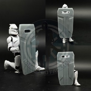 May include: A grey plastic shield with a rectangular shape and a small rectangular opening in the centre. The shield has a raised design on the surface.