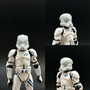 Puede incluir: Una figura de acción de plástico blanco de un soldado clon de Star Wars con un diseño pintado en azul y blanco. La figura tiene un casco con una visera y una pieza de pecho detallada. La figura está en posición de pie con los brazos a los lados.