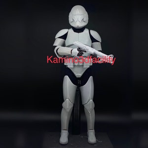May include: A white plastic action figure of a Star Wars clone trooper, holding a blaster rifle. The figure is wearing a white helmet with a red star on the front. The figure is standing on a black base.
