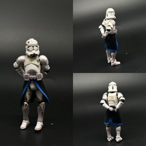 May include: A white and gray Star Wars action figure with a black and blue cape. The figure is standing with its arms crossed and is wearing a helmet.