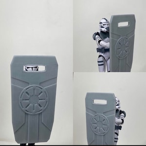 May include: A gray plastic shield with a raised circular design featuring a starburst pattern. The shield has a rectangular shape with a small rectangular opening at the top.