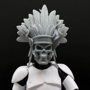 Puede incluir: Una figura gris con rostro de calavera y tocado de plumas, con armadura blanca. La calavera de la figura es detallada, con dientes y cuencas oculares visibles. El tocado presenta grandes plumas estilizadas. La figura está sobre un fondo negro.