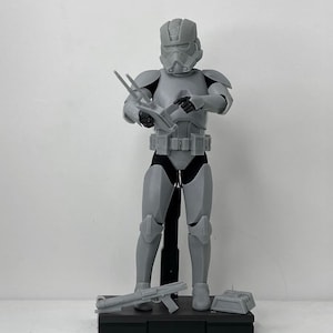Può includere: Una action figure di plastica grigia di un clone trooper di Star Wars con un fucile blaster e un elmetto grigio. La figura è in piedi su una base nera.
