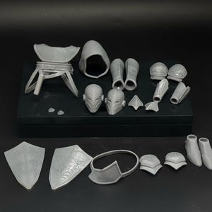 Peut inclure: Ensemble d'éléments d'armure miniatures gris non peints, comprenant un casque, des épaulières, des bottes et des boucliers. Les composants sont disposés sur une surface noire, suggérant un kit de modélisme.