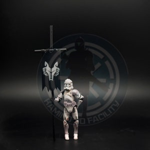 Peut inclure: Figurine d'un soldat clone de Star Wars en armure blanche et grise, tenant une bannière noire avec un emblème de loup. La figurine se dresse sur un fond sombre avec un logo et une silhouette.