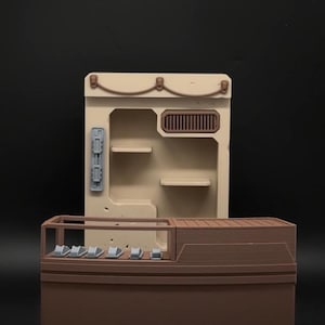 Puede incluir: Maqueta de dos piezas de un interior de tienda, en beige y marrón. La parte trasera tiene estanterías y una rejilla de ventilación, mientras que la parte delantera tiene un mostrador con pequeñas áreas de exhibición. El modelo está sobre un fondo oscuro.