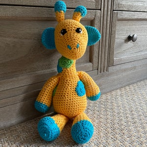 Könnte beinhalten: Ein gehäkeltes Giraffenspielzeug mit gelbem Fell, blauen Ohren und Füßen und grünen Flecken. Die Giraffe sitzt auf einem hellbraunen Teppich.