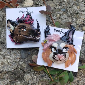 Op de afbeelding: Twee handgemaakte kleikunstwerken met gestileerde dierengezichten. Eén is bruin met zwarte en roze accenten, de andere is tan, grijs en roze. Beide zijn gemonteerd op witte kaarten met het woord "Veneficae" erop.