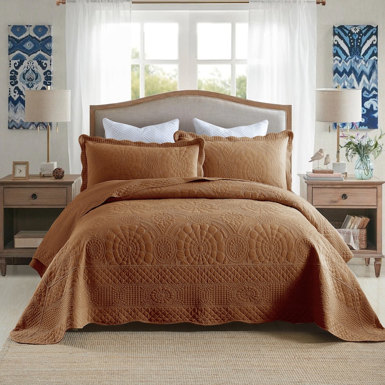 K&ouml;nnte beinhalten: Ein brauner gesteppter Bett&uuml;berwurf mit einem dekorativen Blumenmuster. Der Bett&uuml;berwurf liegt auf einem Bett mit zwei braunen Kissenbez&uuml;gen und zwei wei&szlig;en Kissen.