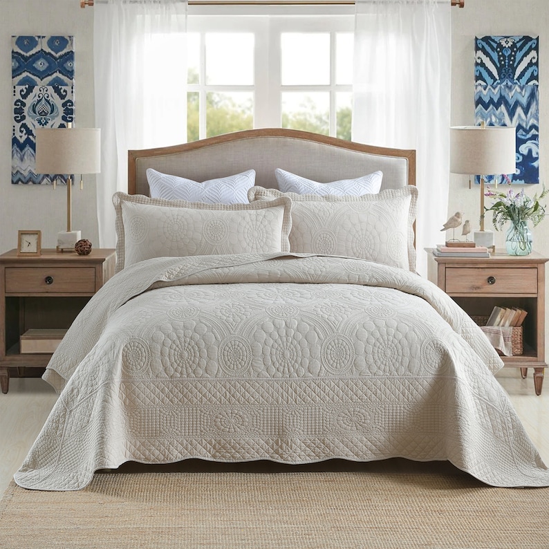 K&ouml;nnte beinhalten: Eine beige gesteppte Bettdecke mit Blumenmuster. Die Bettdecke liegt auf einem Bett mit wei&szlig;en Kissen und einem beigefarbenen Kopfteil. An beiden Seiten des Bettes stehen zwei Lampen auf Nachttischen.