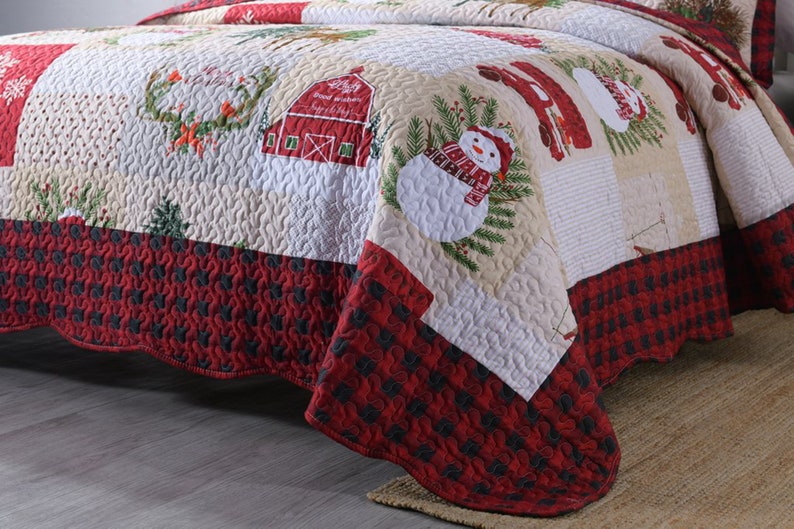 May include: A red and white patchwork quilt with a Christmas theme. The quilt features a snowman, a barn, and a wreath. The quilt has a red and black checkered border.