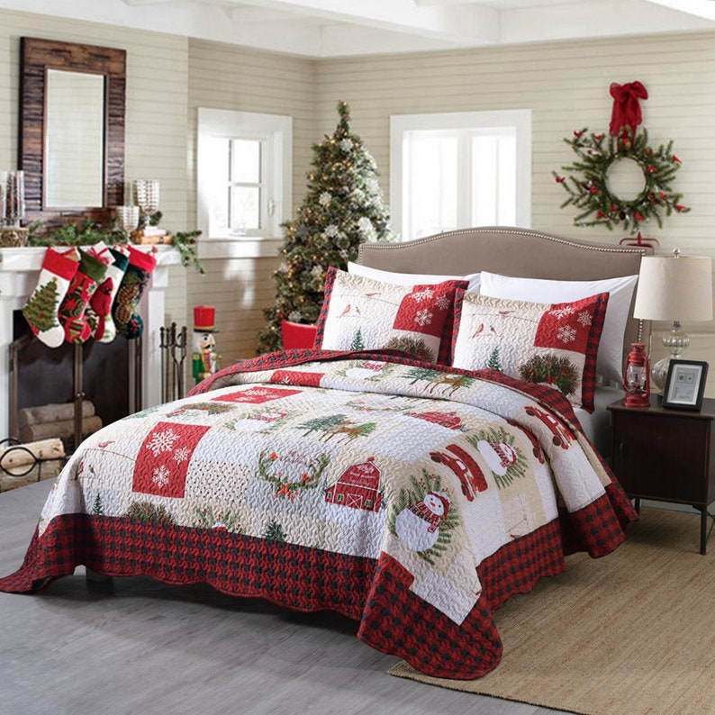 May include: A red and white Christmas themed quilt with a patchwork design featuring snowmen, reindeer, and a farmhouse. The quilt is on a bed with two matching pillowcases.