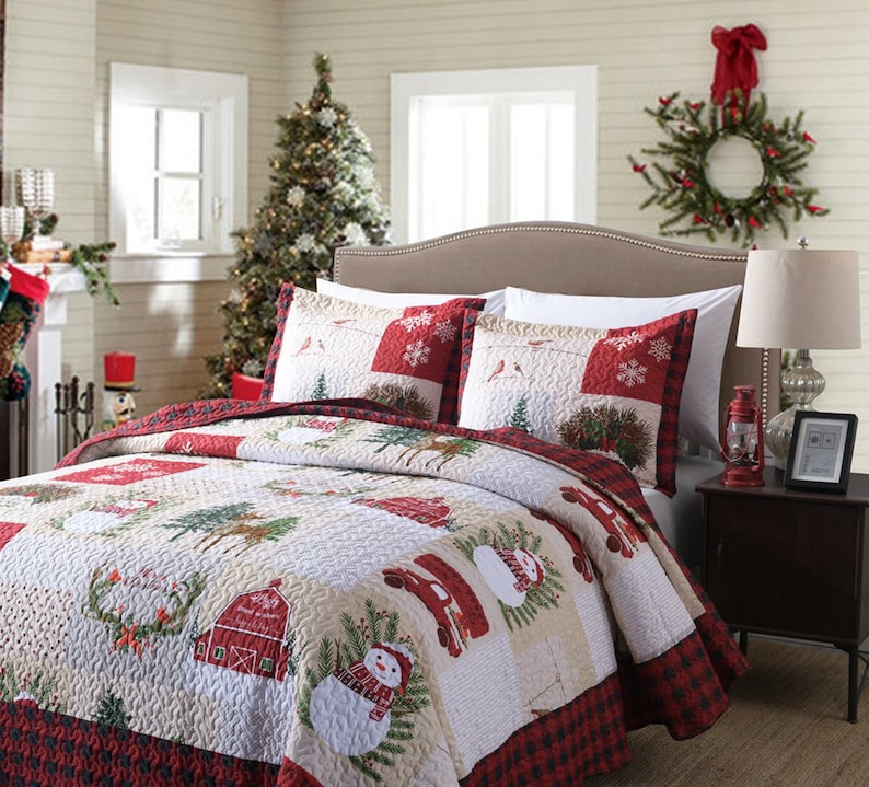May include: A red and white Christmas themed quilt set with a snowman, a red truck, and a barn. The quilt is made of a soft, quilted fabric and features a patchwork design.