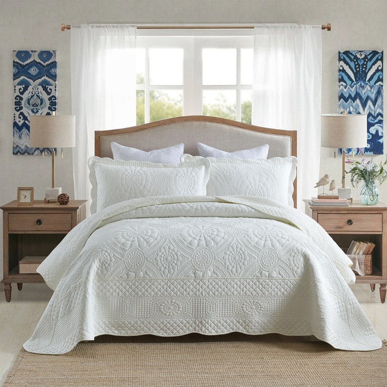 K&ouml;nnte beinhalten: Ein wei&szlig;er gesteppter Bett&uuml;berwurf mit Blumenmuster. Der Bett&uuml;berwurf liegt auf einem Bett mit wei&szlig;en Kissen und einem wei&szlig;en Kopfteil. Das Bett steht in einem Schlafzimmer mit wei&szlig;en Vorh&auml;ngen und einem Nachttisch aus Holz.
