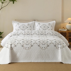 Pode incluir: Uma colcha branca e duas fronhas a condizer com um padrão floral circular repetitivo em tons de cinzento e castanho suaves. O conjunto de cama tem uma textura acolchoada com bordas recortadas, cobrindo uma estrutura de cama neutra.