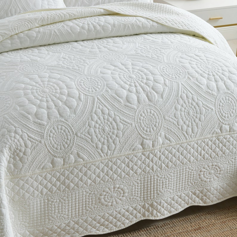 K&ouml;nnte beinhalten: Wei&szlig;e gesteppte Bettdecke mit Blumenmuster. Die Bettdecke hat einen Rautenmuster am unteren Rand.