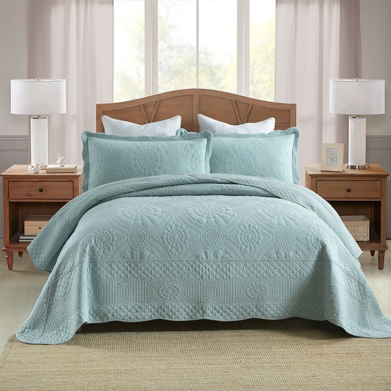 K&ouml;nnte beinhalten: Eine hellblaue gesteppte Bettdecke mit Blumenmuster. Die Bettdecke liegt auf einem Bett mit zwei wei&szlig;en Kissen und einem Kopfteil aus Holz.