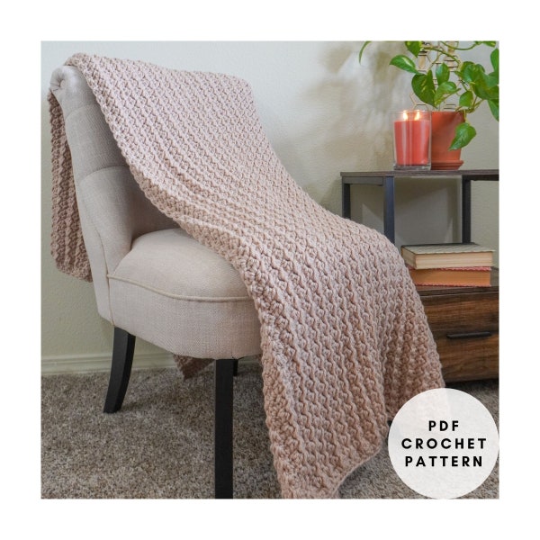 May include: A light brown, textured crochet blanket draped over a beige armchair. The blanket has a repeating pattern and is folded over the chair's back and seat. A small, round graphic with the text "PDF CROCHET PATTERN" is in the lower right corner.