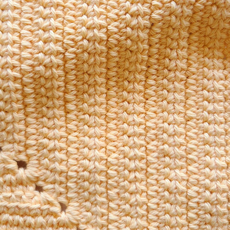 May include: Close-up of a crocheted item in a soft, pale yellow color. The texture is dense and intricate, with visible stitches creating a raised pattern. The yarn appears to be a natural fiber, and the overall effect is warm and inviting.