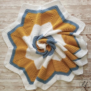 A crocheted baby blanket in white, mustard yellow, and blue. The blanket is folded in a spiral shape and has a starburst pattern.