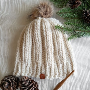 Peut inclure: Bonnet en tricot c&ocirc;tel&eacute; de couleur cr&egrave;me avec un pompon marron duveteux sur le dessus. Le chapeau a une petite &eacute;tiquette en cuir marron. L'arri&egrave;re-plan comprend des aiguilles de pin et des pommes de pin, sugg&eacute;rant un th&egrave;me hivernal. Un crochet est &eacute;galement visible.