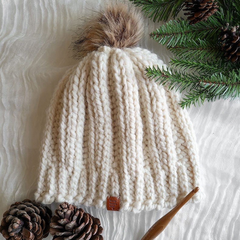 Peut inclure: Bonnet en tricot c&ocirc;tel&eacute; de couleur cr&egrave;me avec un pompon marron duveteux sur le dessus. Le chapeau a une petite &eacute;tiquette en cuir marron. L'arri&egrave;re-plan comprend des aiguilles de pin et des pommes de pin, sugg&eacute;rant un th&egrave;me hivernal. Un crochet est &eacute;galement visible.