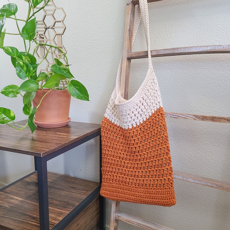 This image shows a finished bag from this crochet tote bag pattern. It is made in orange and cream and is hanging from a blanket ladder. There is also a side table, a plant and a candle.