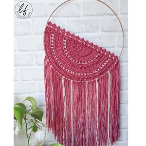 A finished sample of this crochet wall hanging pattern hung on a white brick wall with a plant in the corner. The crochet wall hanging is made in pink and white.
