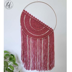 A finished sample of this crochet wall hanging pattern hung on a white wall with a plant in the corner. The crochet wall hanging is made in pink and white.