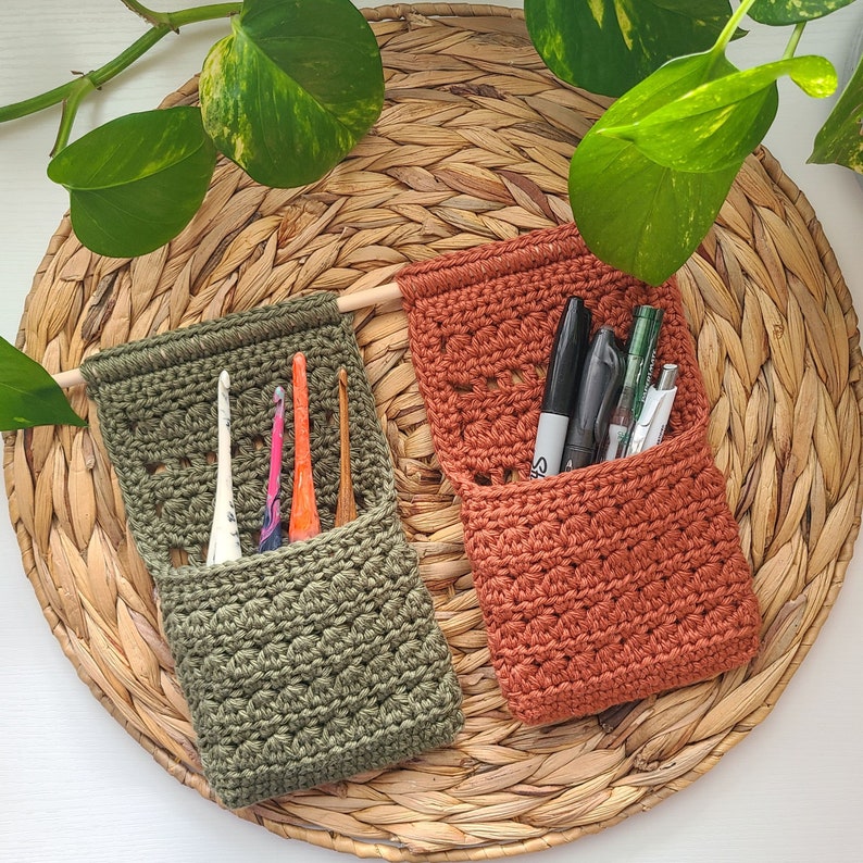 Two handmade crochet wall organizers in olive green and rust orange, each with a wooden dowel for hanging. The olive green organizer holds crochet hooks, while the rust orange one contains pens and markers. The organizers are displayed on a woven wicker tray.