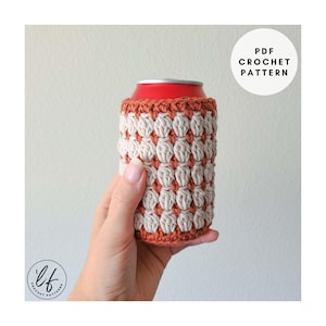 Peut inclure: Un protège-canette crocheté à la main, aux couleurs crème et rouille. Le protège-canette présente un motif de point texturé et est conçu pour s'adapter à une canette de boisson standard. Le texte "PDF Crochet Pattern" est inclus.