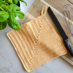 May include: A handmade, square, peach-colored crochet potholder with a loop for hanging. The potholder is placed on a wooden cutting board next to a black knife and a whisk. A green plant is visible in the background.