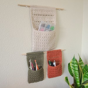 Three crochet hanging baskets made in cream, olive green, and burnt orange. Each crochet basket hangs from a wooden dowel. The cream organizer holds a clear plastic container with colorful pins. The green organizer holds toothbrushes. The orange organizer holds pens.