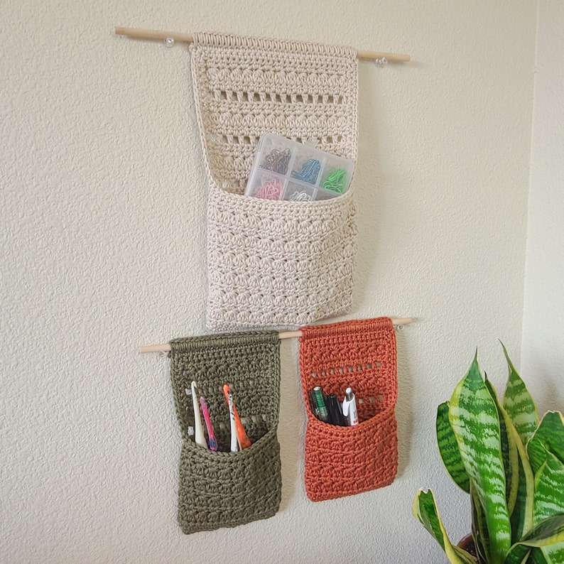 Three crochet hanging baskets made in cream, olive green, and burnt orange. Each crochet basket hangs from a wooden dowel. The cream organizer holds a clear plastic container with colorful pins. The green organizer holds toothbrushes. The orange organizer holds pens.