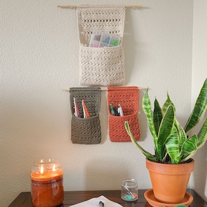 A wall-mounted crochet organizer of crochet hanging baskets in beige, olive green, and rust orange. The organizer has pockets for storage and is suspended from wooden dowels. Sunglasses and eyeglasses are visible in the pockets.