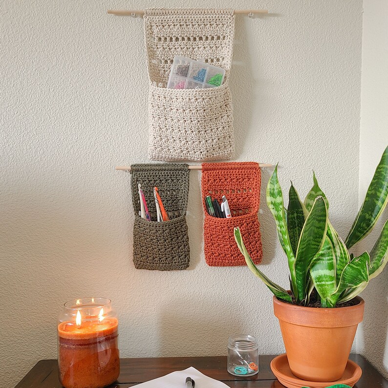 A wall-mounted crochet organizer of crochet hanging baskets in beige, olive green, and rust orange. The organizer has pockets for storage and is suspended from wooden dowels. Sunglasses and eyeglasses are visible in the pockets.