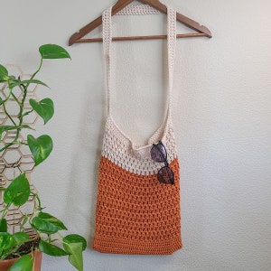 This image shows a finished bag from this crochet tote bag pattern. It is made in orange and cream and is hanging from a blanket ladder. There is also a side table, a plant and a candle.