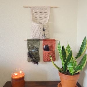 A wall-mounted organizer with three crocheted pockets in beige, olive green, and rust orange. The pockets hang from wooden dowels. Sunglasses and other items are visible.