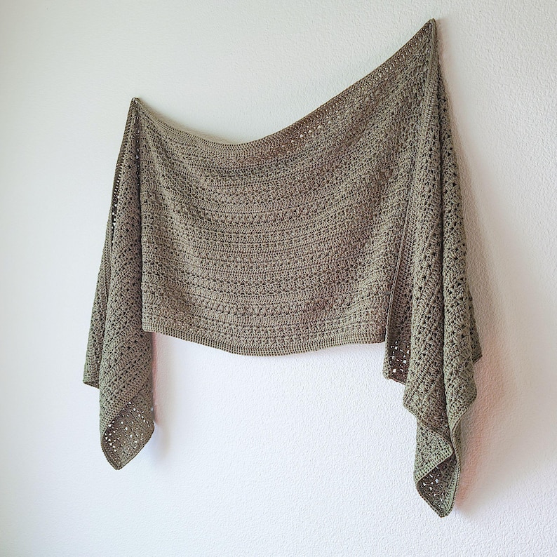A sage green crochet shawl with a textured pattern and openwork detailing. The shawl is draped against a white wall.