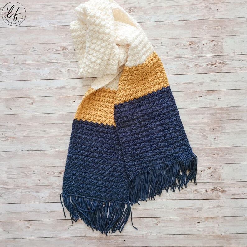 This image shows a sample made from this scarf crochet pattern. The scarf is made in blue, yellow and white and laid flat on a faux wood background.