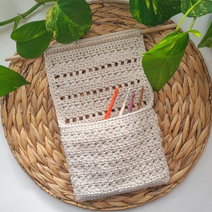 A beige crochet hanging basket with a wooden dowel for hanging. The organizer has a pocket containing several crochet hooks in various colors. The piece is set against a woven background with green foliage.