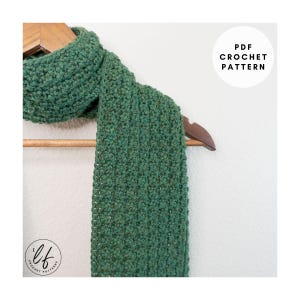 Op de afbeelding: Een groene gehaakte sjaal hangt aan een houten kleerhanger. De sjaal heeft een textuur met ribbels. De tekst "PDF CROCHET PATTERN" staat in een witte cirkel. De sjaal is een handgemaakt item.